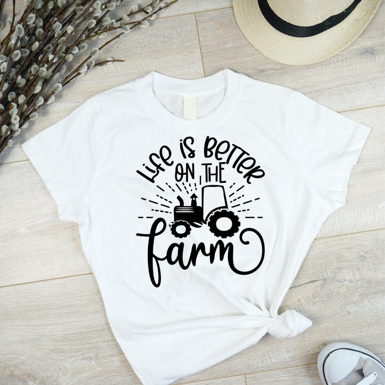 May include: White t-shirt with black text that reads "Life is better on the farm" with a black silhouette of a tractor in the center.