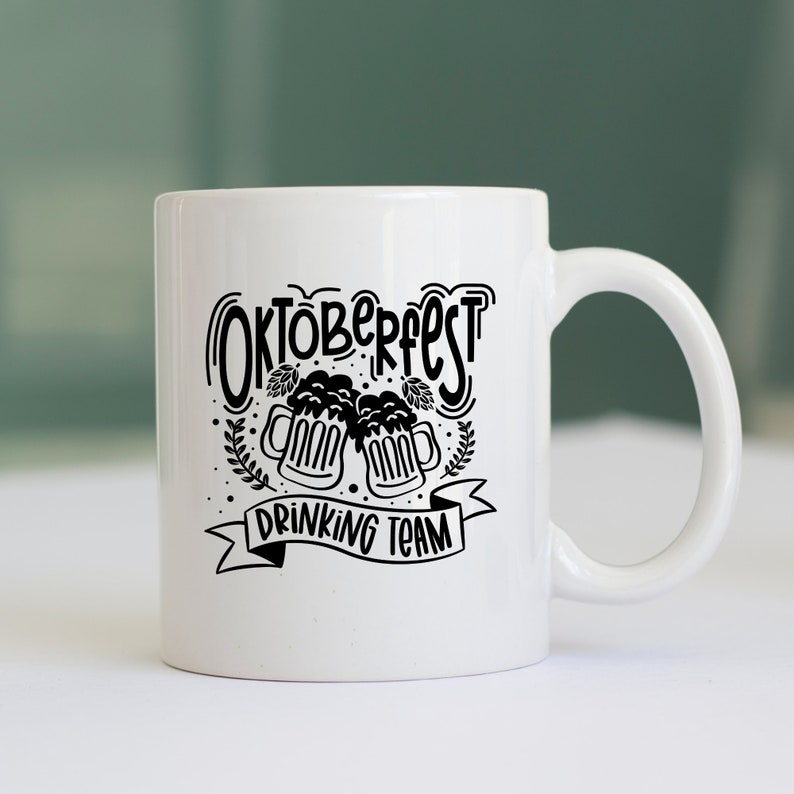 May include: White ceramic mug with a black design that says "Oktoberfest Drinking Team" with two beer mugs clinking together.