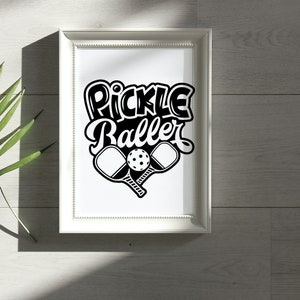 Pode incluir: Uma impress&atilde;o emoldurada branca com o texto "Pickle Baller" em letras estilizadas pretas. Duas raquetes de pickleball pretas e uma bola de pickleball branca s&atilde;o mostradas abaixo do texto.
