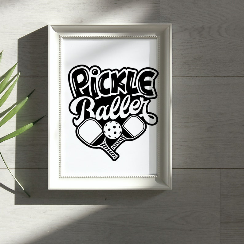 Pode incluir: Uma impress&atilde;o emoldurada branca com o texto "Pickle Baller" em letras estilizadas pretas. Duas raquetes de pickleball pretas e uma bola de pickleball branca s&atilde;o mostradas abaixo do texto.