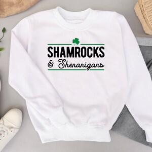 Op de afbeelding: Witte trui met een groene klaver en de tekst "Shamrocks & Shenanigans" in zwart.