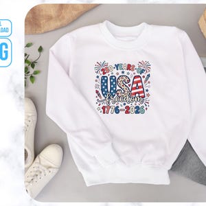 May include: A white sweatshirt featuring a vibrant graphic commemorating 250 years of USA freedom. The design incorporates the text "250 Years of USA Freedom 1776-2026" in red, white, and blue, with star and firework motifs. The sweatshirt is shown on a neutral background.