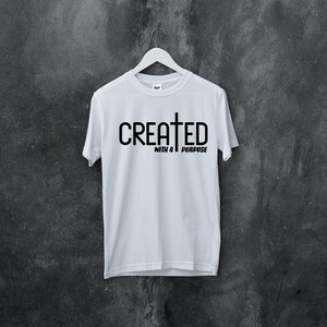 以下が含まれることがあります： 黒い文字で「CREATED WITH A PURPOSE」と十字架のシンボルが書かれた白いTシャツです。