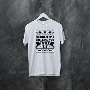 Puede incluir: Camiseta blanca con un dise&ntilde;o negro de hockey. El dise&ntilde;o presenta dos jugadores de hockey y el texto "Making a fist checking you twice".