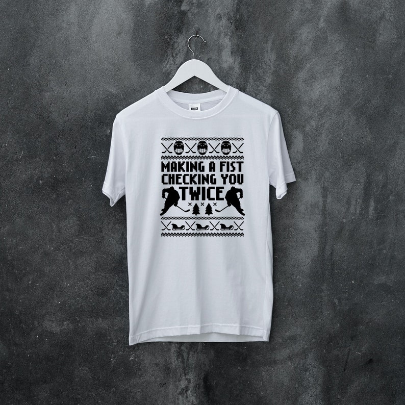 Puede incluir: Camiseta blanca con un dise&ntilde;o negro de hockey. El dise&ntilde;o presenta dos jugadores de hockey y el texto "Making a fist checking you twice".
