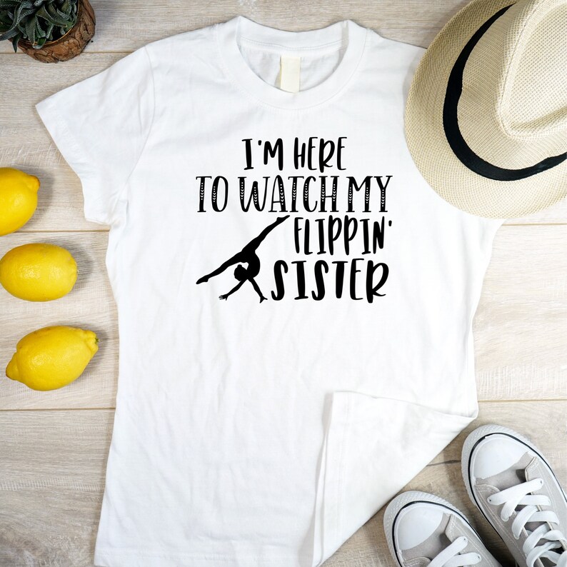 May include: White t-shirt with black text that reads "I'm here to watch my flippin' sister" and a silhouette of a gymnast doing a back handspring.