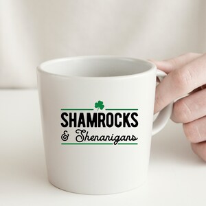 Op de afbeelding: Witte keramische mok met een zwart en groen klavertjevier ontwerp en de tekst "Shamrocks & Shenanigans".