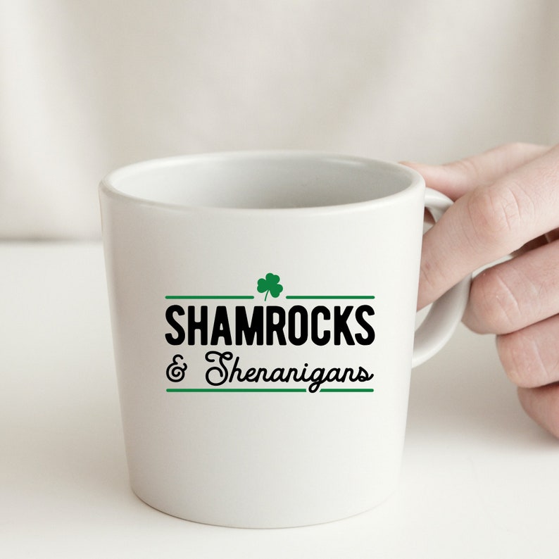 Op de afbeelding: Witte keramische mok met een zwart en groen klavertjevier ontwerp en de tekst "Shamrocks & Shenanigans".