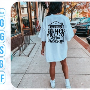 Op de afbeelding: Wit T-shirt met een zwart grafisch ontwerp van een huis met de tekst "We said yes to the address".