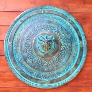 May include: A round, teal-colored metal shield with intricate carvings and a raised, bronze-colored, dragon-like creature in the center.
