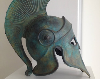 May include: A bronze Greek warrior helmet with a green patina finish. The helmet has a tall, crested plume and a face mask.