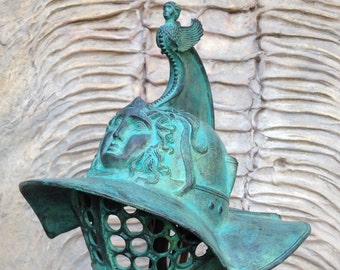 May include: A green bronze gladiator helmet with a face mask and a winged figure on top.