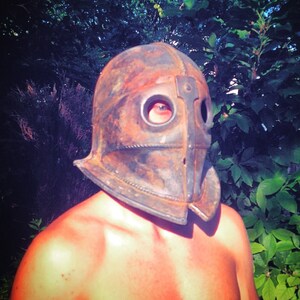 Könnte beinhalten: Eine Person trägt einen rostigen Metallhelm mit einem einzigen Augenloch. Der Helm ist in Form einer abgerundeten Kuppel mit einer spitzen Vorderseite.