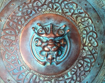May include: A circular copper and turquoise metal plate with an intricate design of Celtic knots and a central embossed image of a mythical creature with a fierce expression.