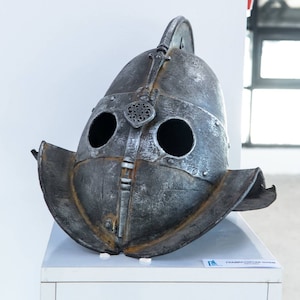 Puede incluir: Un casco metálico envejecido con visera y agujeros para los ojos, probablemente para recreaciones históricas o exhibiciones. El casco tiene una parte superior redondeada con una cresta decorativa y una construcción remachada. Se encuentra sobre una superficie blanca.