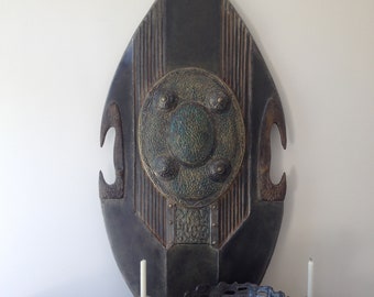 May include: A dark wooden shield with a metal center piece and decorative accents. The shield is hanging on a white wall above a wooden table with two candlesticks and a bowl of fruit.