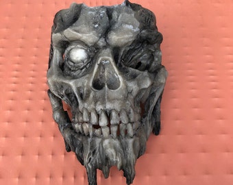 May include: A detailed, gray and white skull mask with a single, white eye. The mask is made of a textured material and has a dripping effect around the jawline.