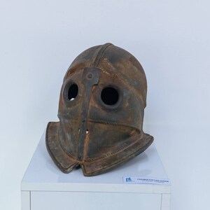 Puede incluir: Un casco de metal oxidado de color marr&oacute;n con dos agujeros para los ojos y una barbada puntiaguda. El casco est&aacute; expuesto en un pedestal blanco.