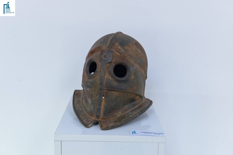 Puede incluir: Un casco de metal oxidado de color marr&oacute;n con dos agujeros para los ojos y una barbada puntiaguda. El casco est&aacute; expuesto en un pedestal blanco.