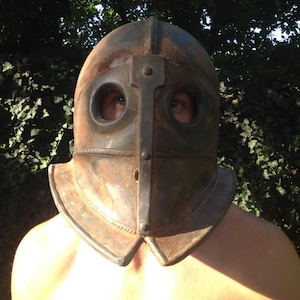 Puede incluir: Un casco de metal oxidado con agujeros para los ojos y una parte superior puntiaguda. El casco lo lleva una persona con los hombros descubiertos.