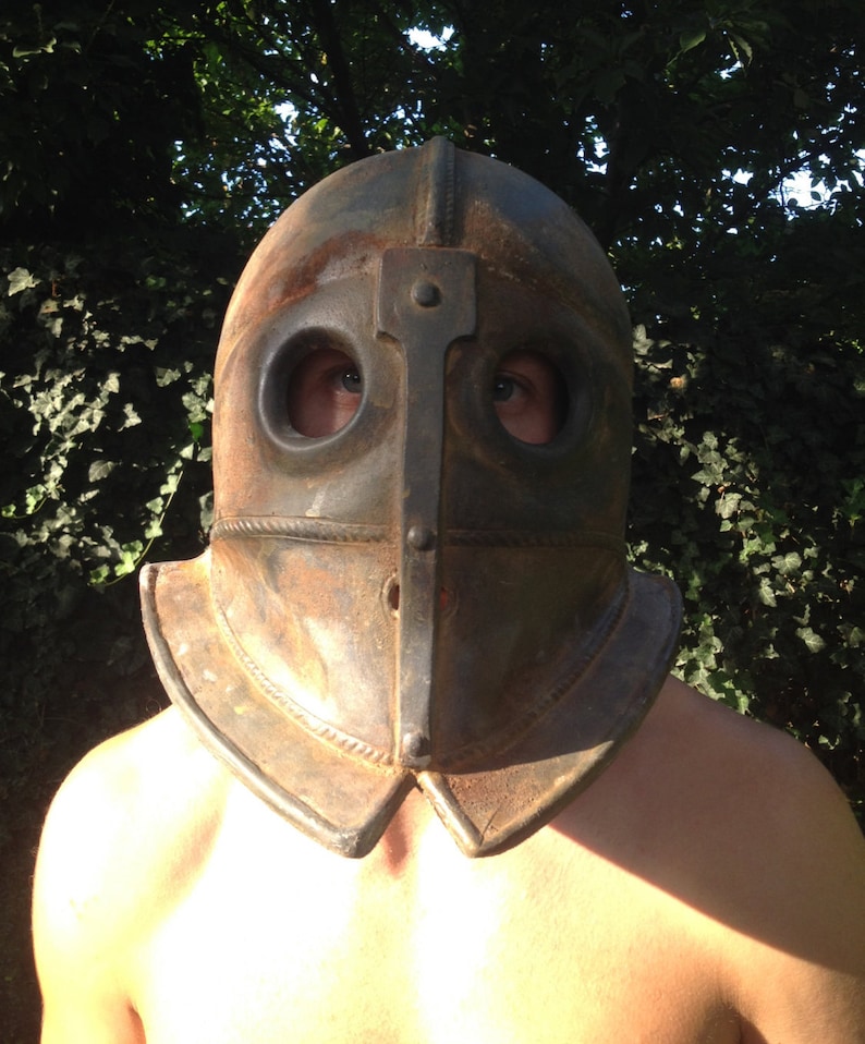 Puede incluir: Un casco de metal oxidado con agujeros para los ojos y una parte superior puntiaguda. El casco lo lleva una persona con los hombros descubiertos.