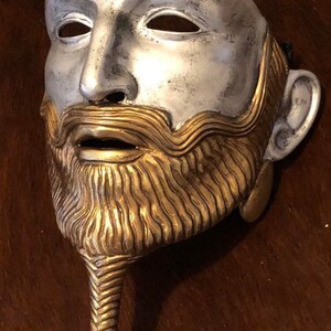 May include: A silver and gold mask with a detailed beard and mustache. The mask has a long, braided beard that ends in a knot.