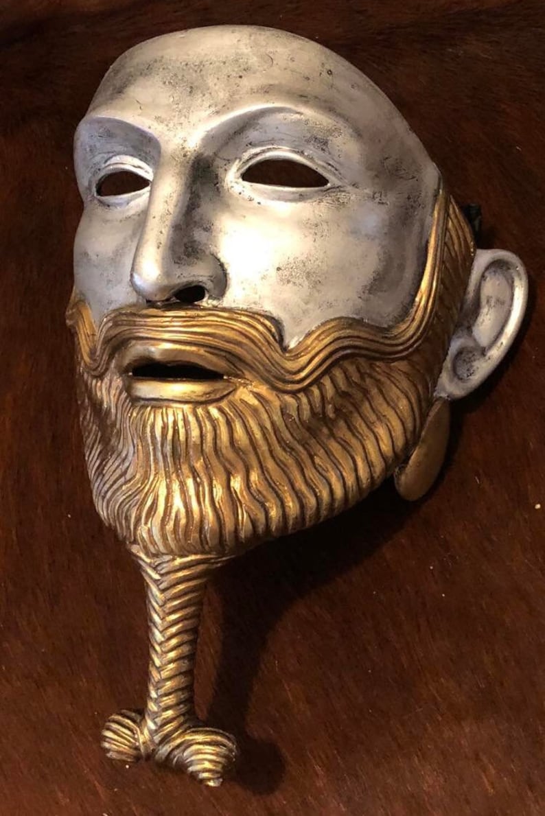 May include: A silver and gold mask with a detailed beard and mustache. The mask has a long, braided beard that ends in a knot.