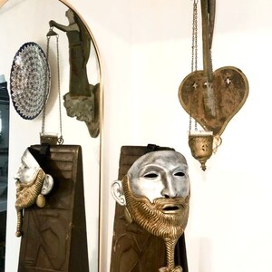 May include: Two silver and gold masks with beards on wooden panels. The masks have a realistic human face with a silver face and a gold beard. The masks are hanging on the wall.