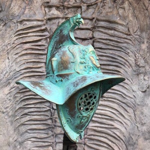 May include: A green metal gladiator helmet with a bird design on the top. The helmet has a face mask with a series of holes. The helmet is on a stand.