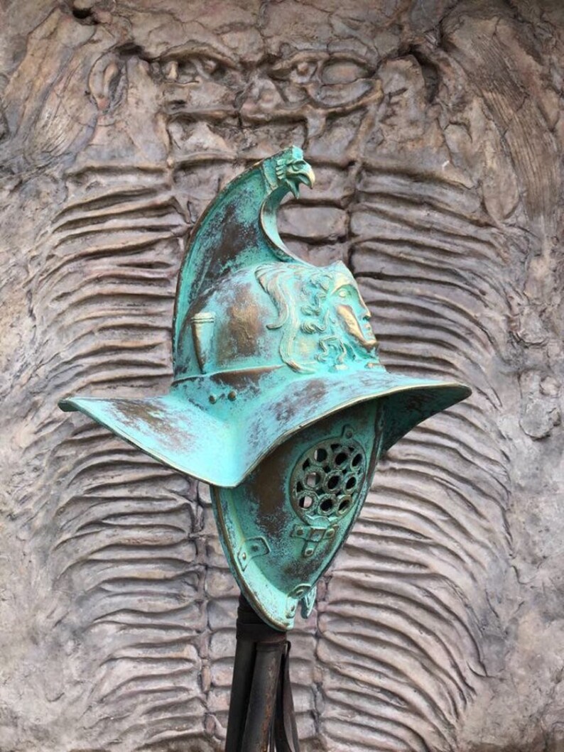 May include: A green metal gladiator helmet with a bird design on the top. The helmet has a face mask with a series of holes. The helmet is on a stand.
