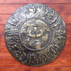 Pode incluir: Um escudo redondo de cor bronze com um desenho em relevo de uma carruagem puxada por dois cavalos e uma figura em pé na carruagem. O escudo é rodeado por uma borda de figuras em relevo.