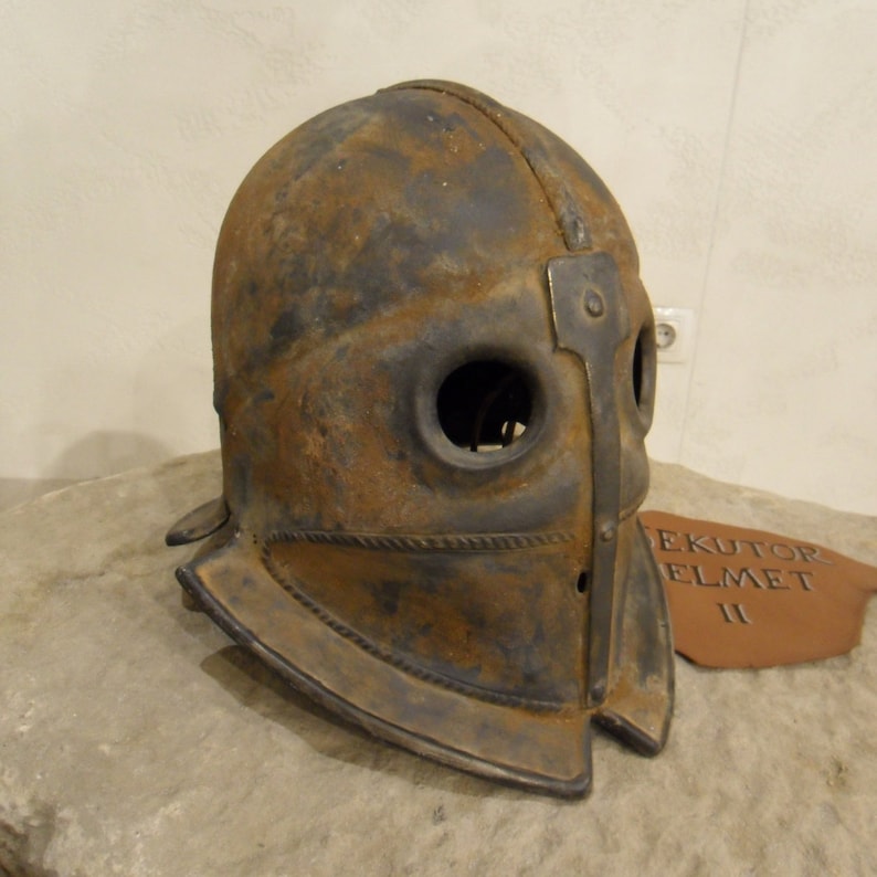 Puede incluir: Un casco de metal oxidado con visera y agujeros para los ojos. El casco est&aacute; etiquetado como "EKUTOR ELMET II".