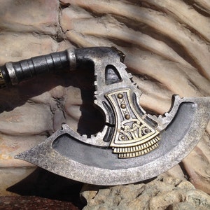 May include: A silver and gold axe with a detailed design on the blade. The axe has a black handle with a gold and silver decorative band.