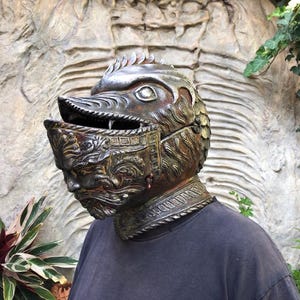 Op de afbeelding: Een gedetailleerde helm in antieke stijl met een vogelachtig ontwerp, met een scharnierend vizier en ingewikkelde gezichtsdetails. De helm is voornamelijk bronskleurig met donkere accenten, gedragen over een donkergrijs shirt.