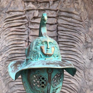 May include: A green bronze gladiator helmet with a face sculpted on the top. The helmet has a large brim and a face mask with a grid pattern.