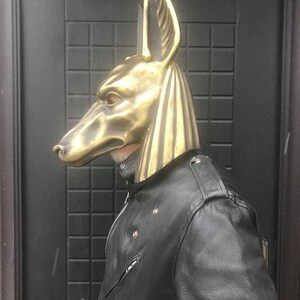 Puede incluir: Una m&aacute;scara de Anubis de color dorado con un dise&ntilde;o detallado. La m&aacute;scara la lleva puesta una persona que lleva una chaqueta de cuero negra.