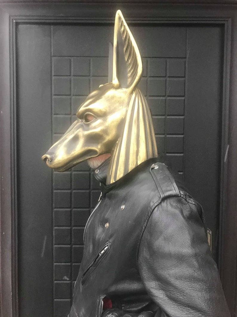 Puede incluir: Una m&aacute;scara de Anubis de color dorado con un dise&ntilde;o detallado. La m&aacute;scara la lleva puesta una persona que lleva una chaqueta de cuero negra.