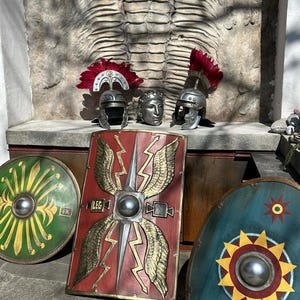 Pode incluir: Uma coleção de capacetes e escudos em estilo romano. Os capacetes têm plumas vermelhas. Os escudos são redondos e retangulares, com vários desenhos, incluindo asas, raios e padrões solares. A paleta de cores inclui vermelho, verde, dourado e azul.