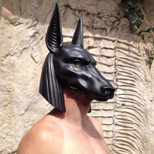 May include: A black Anubis mask with detailed features, including ears, a snout, and eyes. The mask is made of a smooth, hard material and is worn by a person.