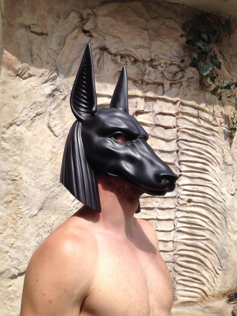 May include: A black Anubis mask with detailed features, including ears, a snout, and eyes. The mask is made of a smooth, hard material and is worn by a person.