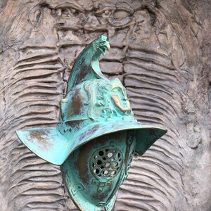 May include: A green metal gladiator helmet with a bird design on the top. The helmet has a face mask with a grill design and a large brim.