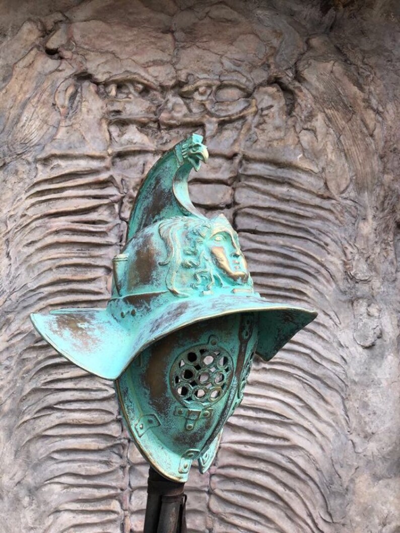 May include: A green metal gladiator helmet with a bird design on the top. The helmet has a face mask with a grill design and a large brim.