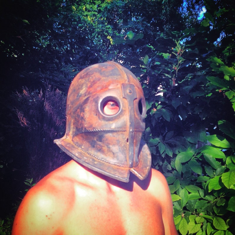 Puede incluir: Una persona que lleva un casco de metal marr&oacute;n oxidado con un solo agujero para el ojo. El casco tiene una protecci&oacute;n de nariz puntiaguda y una superficie texturizada.