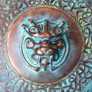 May include: A circular metal shield with a detailed design of swirling patterns and a central embossed image of a mythical creature with a fierce expression. The shield is painted in shades of teal and copper.