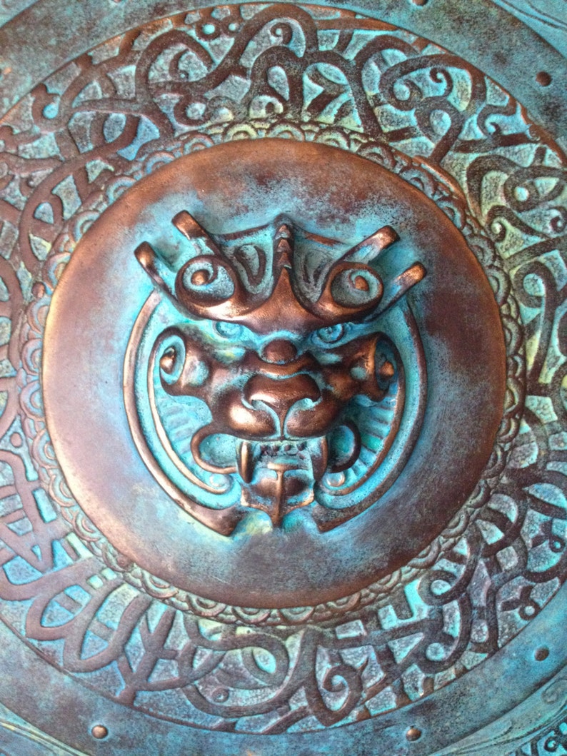May include: A circular metal shield with a detailed design of swirling patterns and a central embossed image of a mythical creature with a fierce expression. The shield is painted in shades of teal and copper.