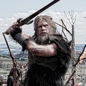 May include: A bearded man in a fur-lined tunic holds a sword in a fighting stance. He is standing in a field with a mountain range in the background.