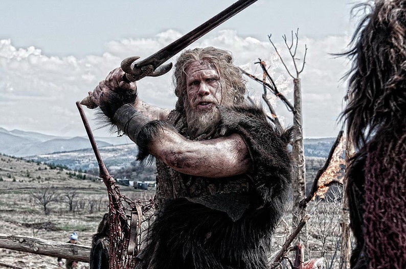 May include: A bearded man in a fur-lined tunic holds a sword in a fighting stance. He is standing in a field with a mountain range in the background.