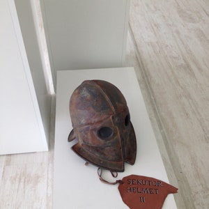 Puede incluir: Un casco de cuero marr&oacute;n con una m&aacute;scara facial de metal y una etiqueta de cuero marr&oacute;n que dice "Sekutor Helmet II".