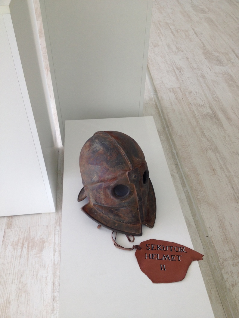 Puede incluir: Un casco de cuero marr&oacute;n con una m&aacute;scara facial de metal y una etiqueta de cuero marr&oacute;n que dice "Sekutor Helmet II".