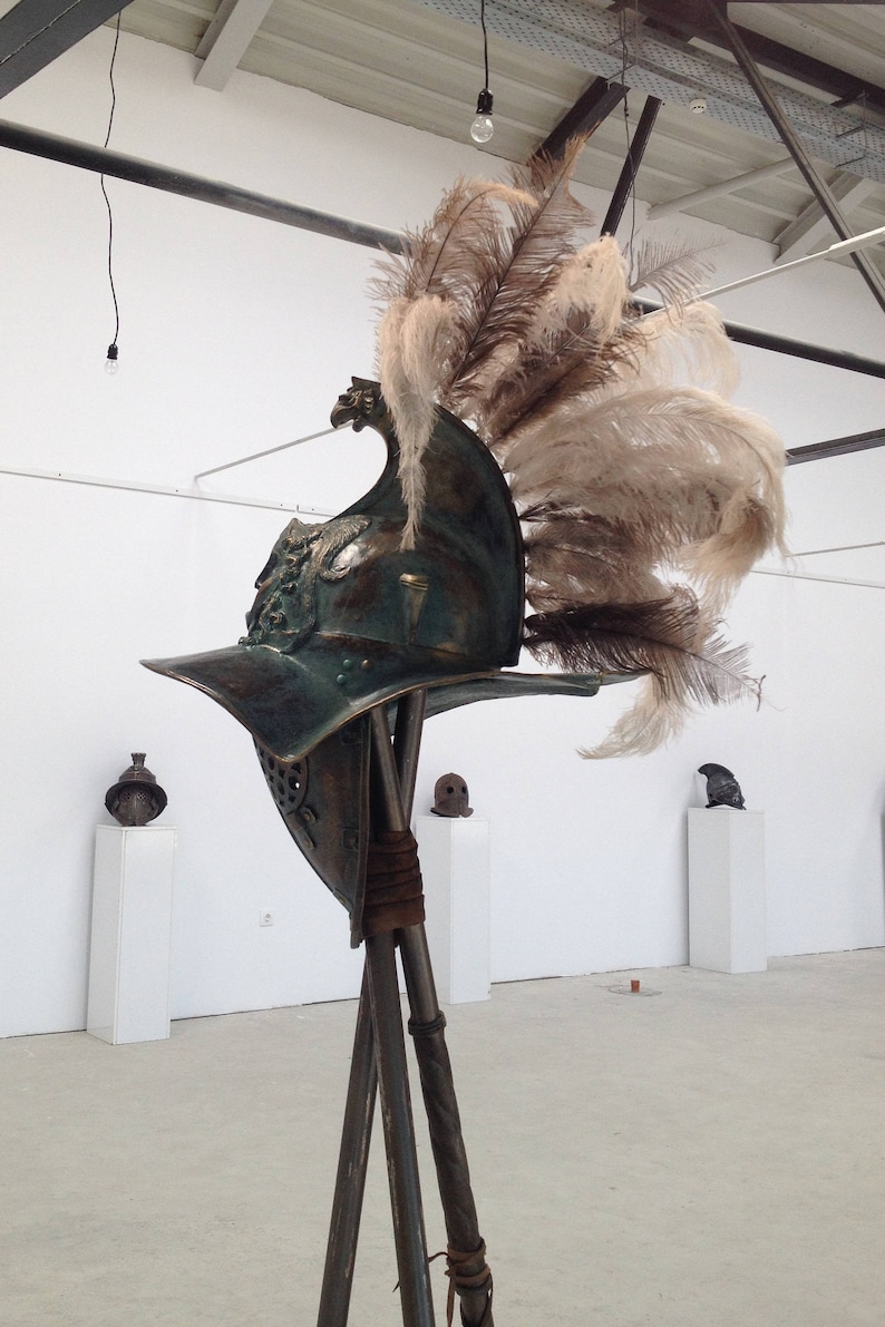 Może przedstawiać: Brązowy hełm gladiatora z pi&oacute;ropuszem z pi&oacute;r na szczycie, zamontowany na drewnianym stojaku. Hełm jest ozdobiony misternymi detalami i ma wizjer.